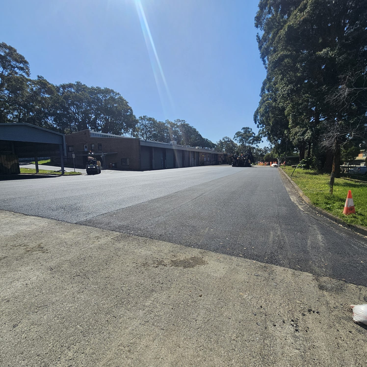 View of a newly paved, wide asphalt road with a row of industrial buildings on the left and trees lining both sides. A traffic cone is visible in the foreground under a clear blue sky.