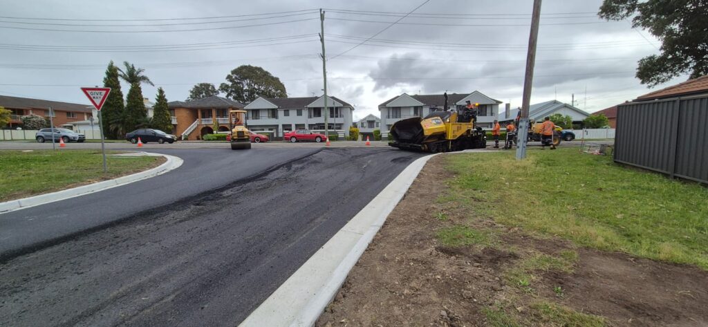 A road construction site with fresh asphalt being laid by a yellow paving machine. Workers are visible, and a steamroller is compacting the surface. Residential houses and overcast skies are in the background. Some traffic cones are on the site.