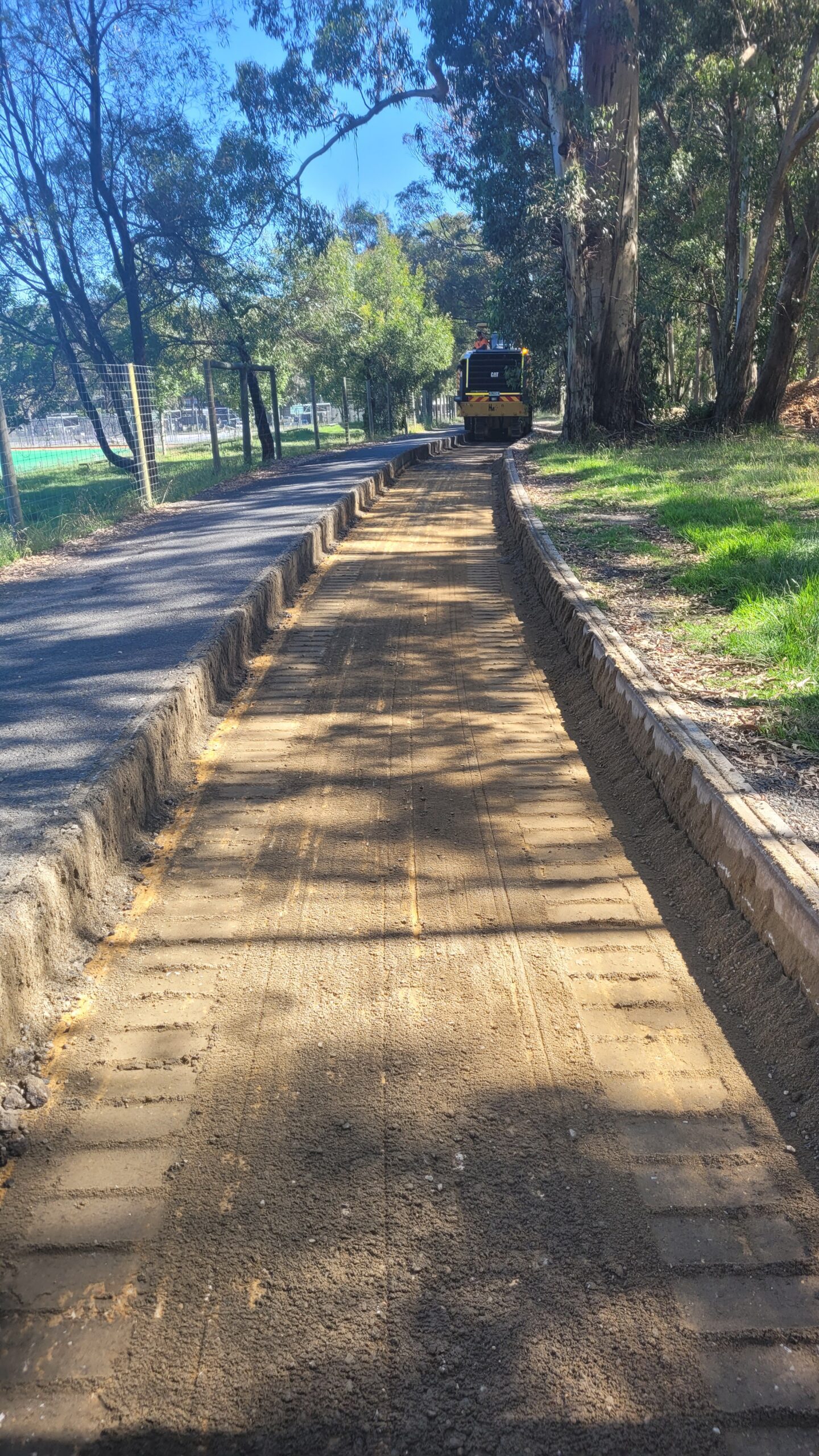 A newly constructed path in a park, bordered by trees and a grassy area, with a construction vehicle visible in the distance. The path is made of compacted dirt and is next to an asphalt walkway.