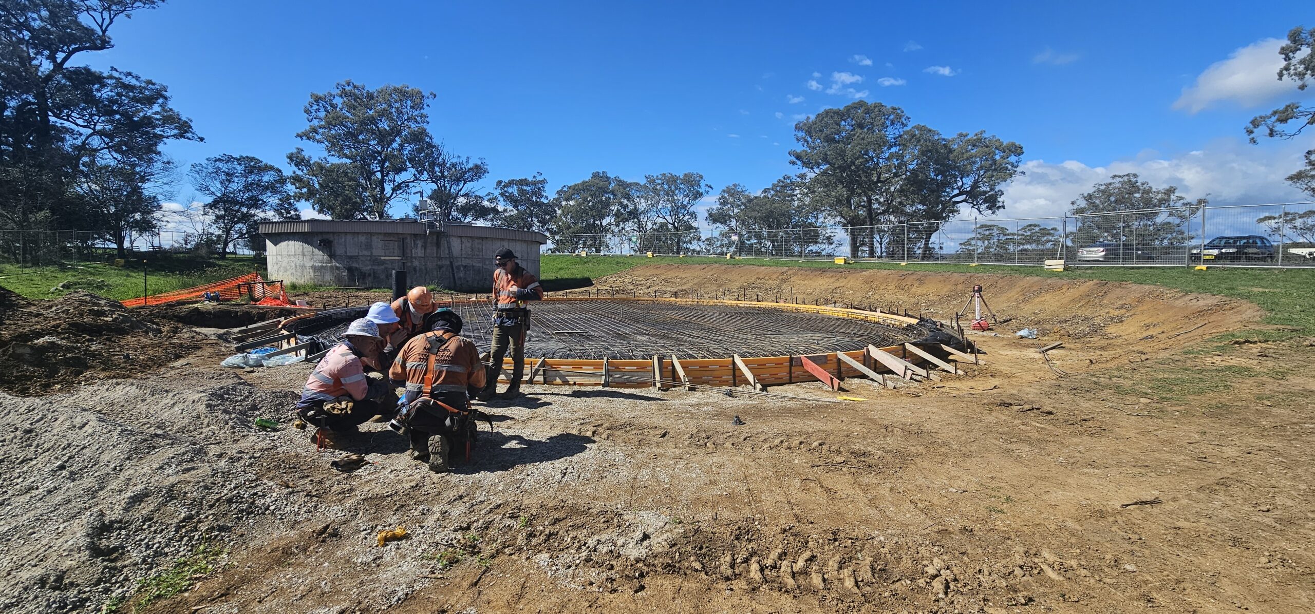 A construction site with a circular foundation in progress, surrounded by dirt and trees. Several workers are gathered, examining plans or equipment. A partially constructed building and more trees are visible in the background under a clear blue sky.