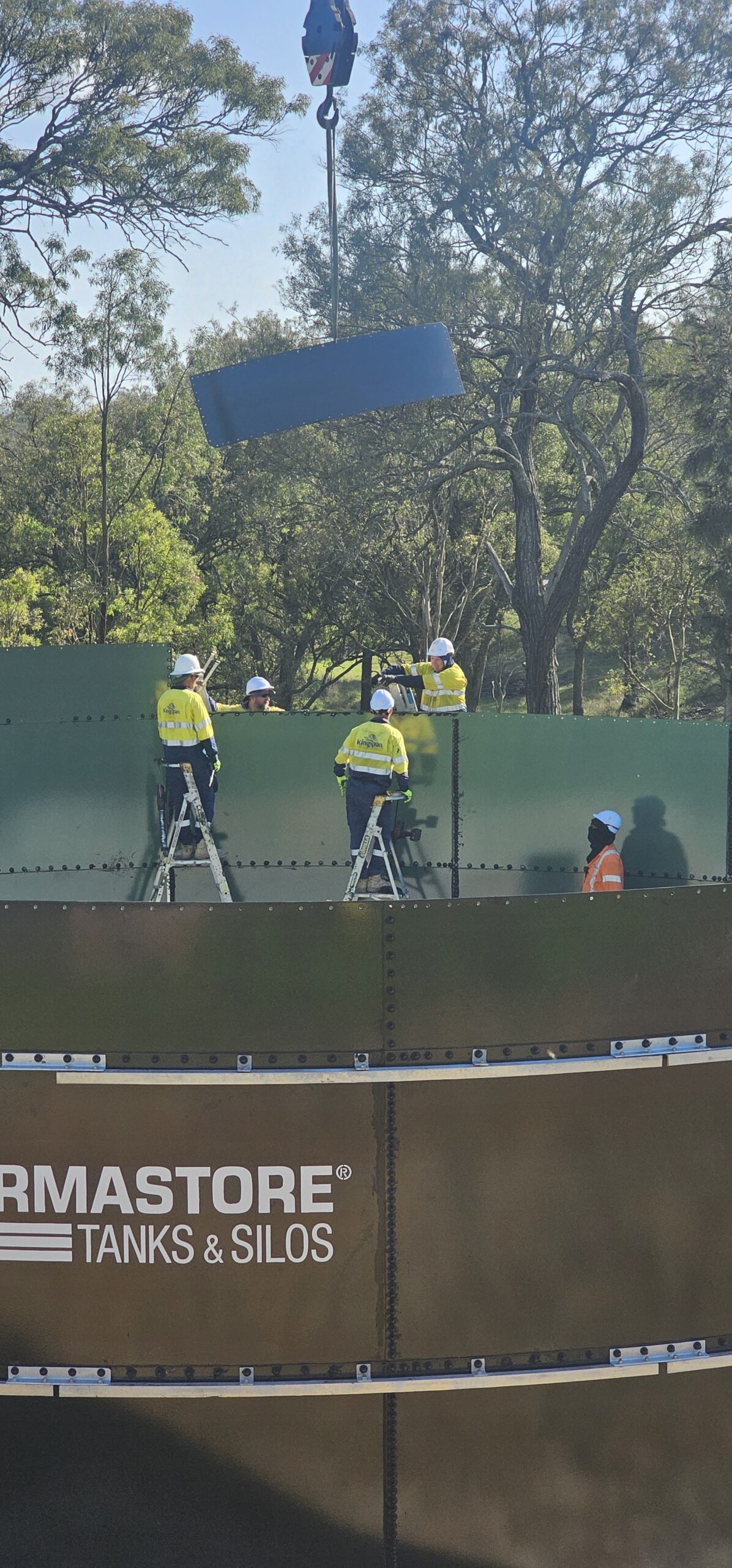 Workers in yellow safety gear assemble a large green tank using ladders and a crane to position a panel. The scene is outdoors with trees in the background.