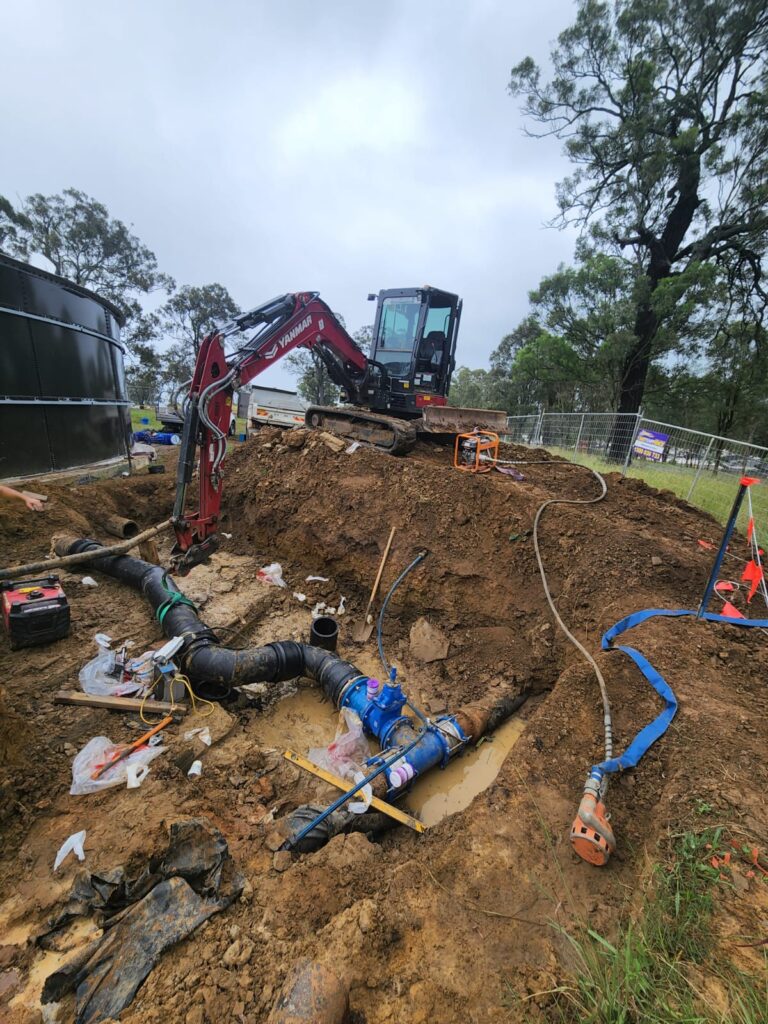 An excavator is positioned on a hillside above a construction site with exposed pipes and water fittings. Muddy water is pooled around the area. Trees and a fenced boundary are visible in the background under an overcast sky.