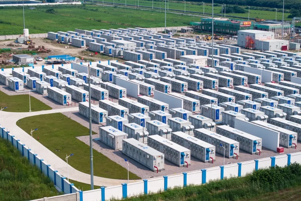 Aerial view of a large battery storage facility with numerous white containers arranged in rows on green grass. There are paths between the rows, light poles, and nearby buildings surrounded by a fence.