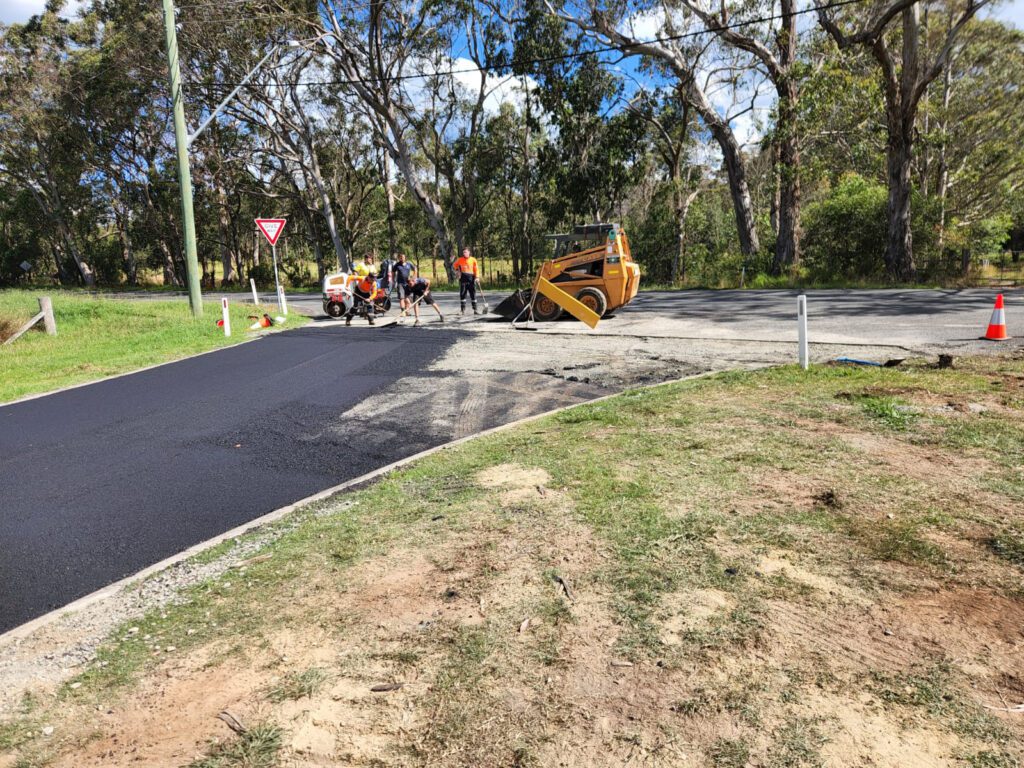 Road construction scene with workers laying asphalt on a rural road. Visible are a steamroller, traffic cones, and trees in the background. A yield sign stands at the intersection.