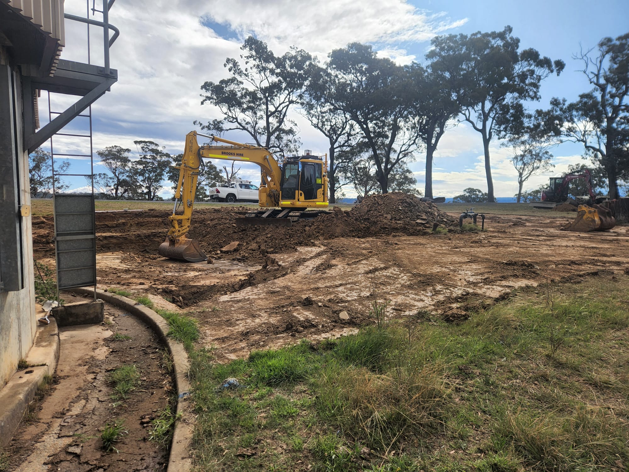 A yellow excavator sits on a construction site, clearing soil and dirt under a partly cloudy sky. Trees and a parked vehicle are visible in the background. A building's edge is on the left, and grass grows along the perimeter of the site.