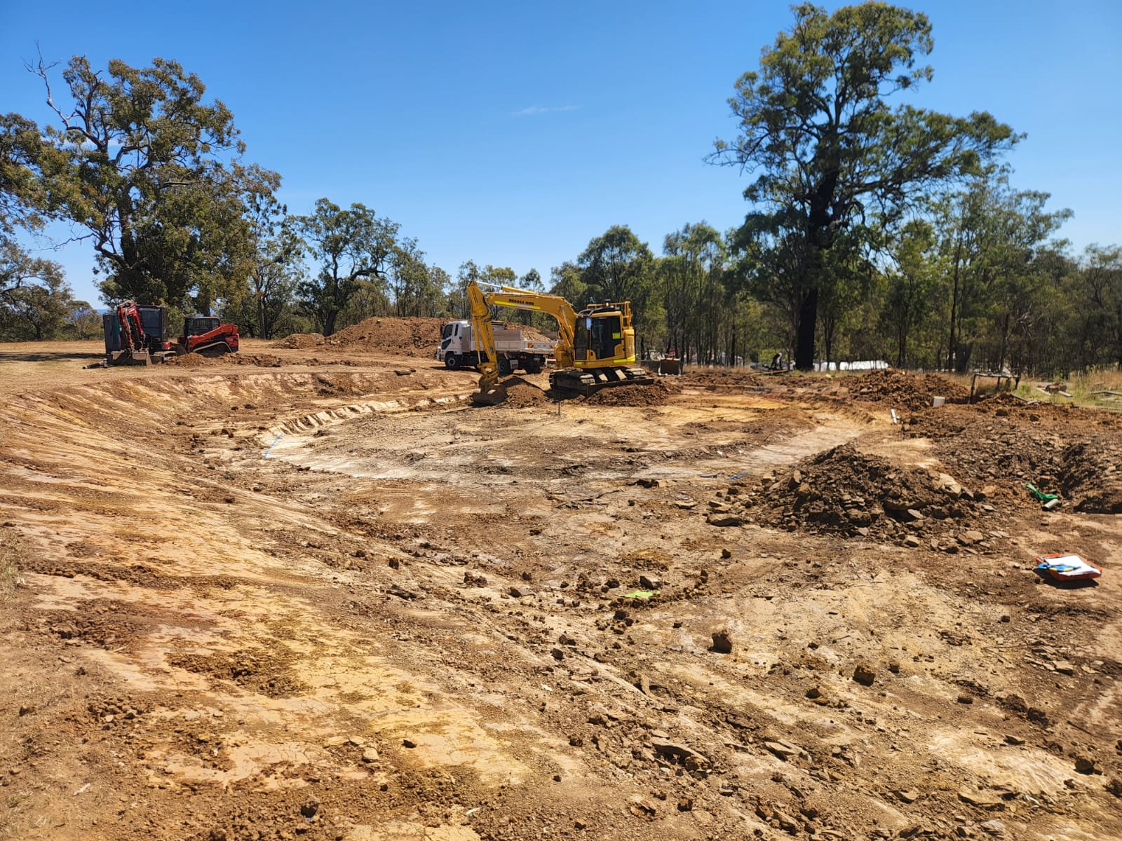 A construction site with a yellow excavator on a large, cleared dirt area surrounded by a few trees. The sky is clear and blue, and there are some machinery and dirt piles in the background.