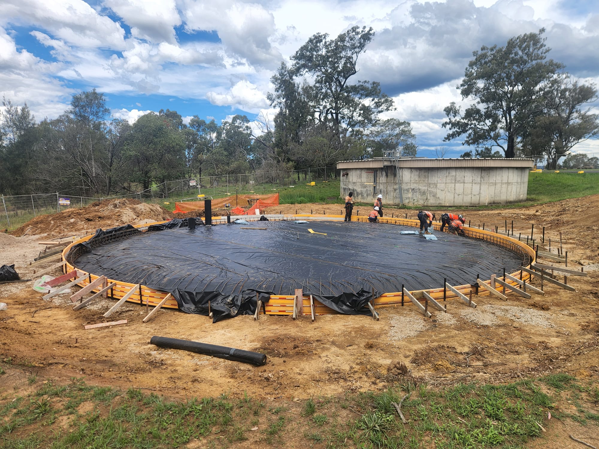 Construction site with a large circular foundation covered in black plastic sheeting. Workers in helmets and safety gear are inspecting the area. Trees and a small building are visible in the background under a partly cloudy sky.