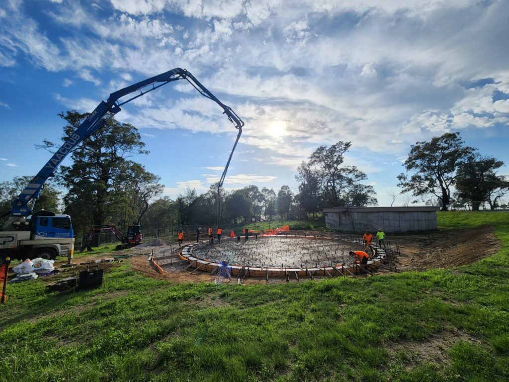 A construction site with workers pouring concrete into a circular foundation using a concrete pump truck. Surrounded by green grass and trees under a partly cloudy blue sky. Safety cones and barricades are placed around the site.