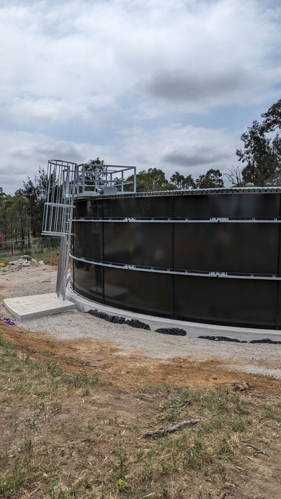 Large cylindrical black water tank with a metal ladder on its side, situated on a concrete base. The surrounding area is grassy with some trees and a partially cloudy sky in the background.