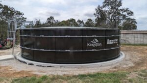 Large cylindrical water tank labeled "Kingspan" and "Permastore" sits on a concrete base. The tank is surrounded by grass and trees under a cloudy sky. A metal ladder leads to the top of the tank.