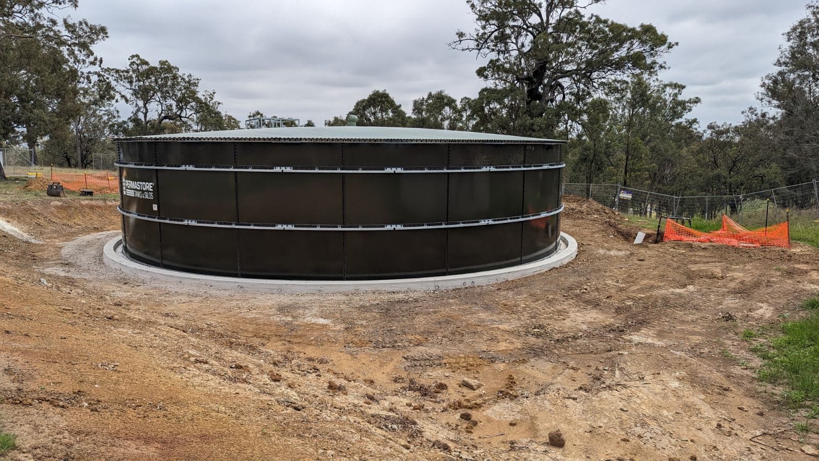 A large, circular black water tank sits on a cleared patch of dirt surrounded by trees. There's orange safety fencing to the right and a gray, cloudy sky above.