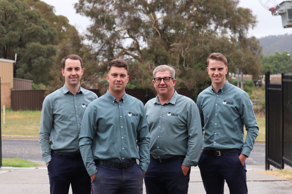 Four men wearing matching blue button-up shirts with a logo stand outdoors. They are posing together in front of trees and a fenced area. The sky is overcast.