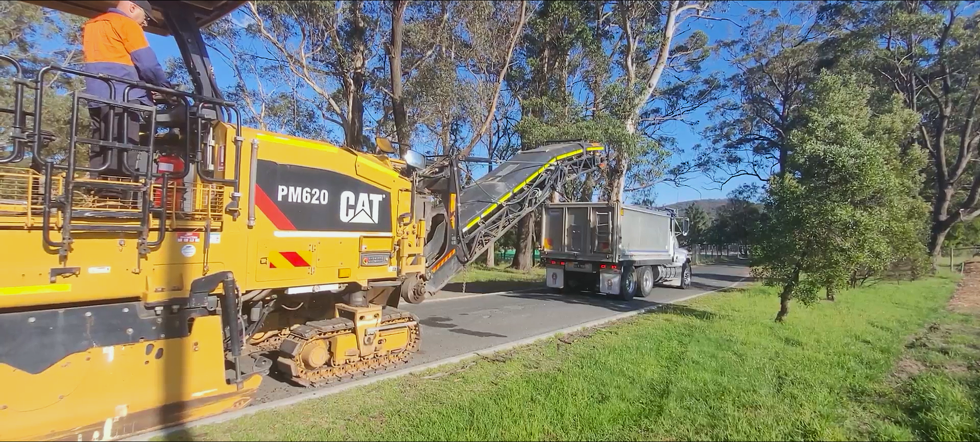 A yellow CAT PM620 road milling machine transfers asphalt to a gray truck on a tree-lined road under a clear blue sky. A grassy area is visible in the foreground.