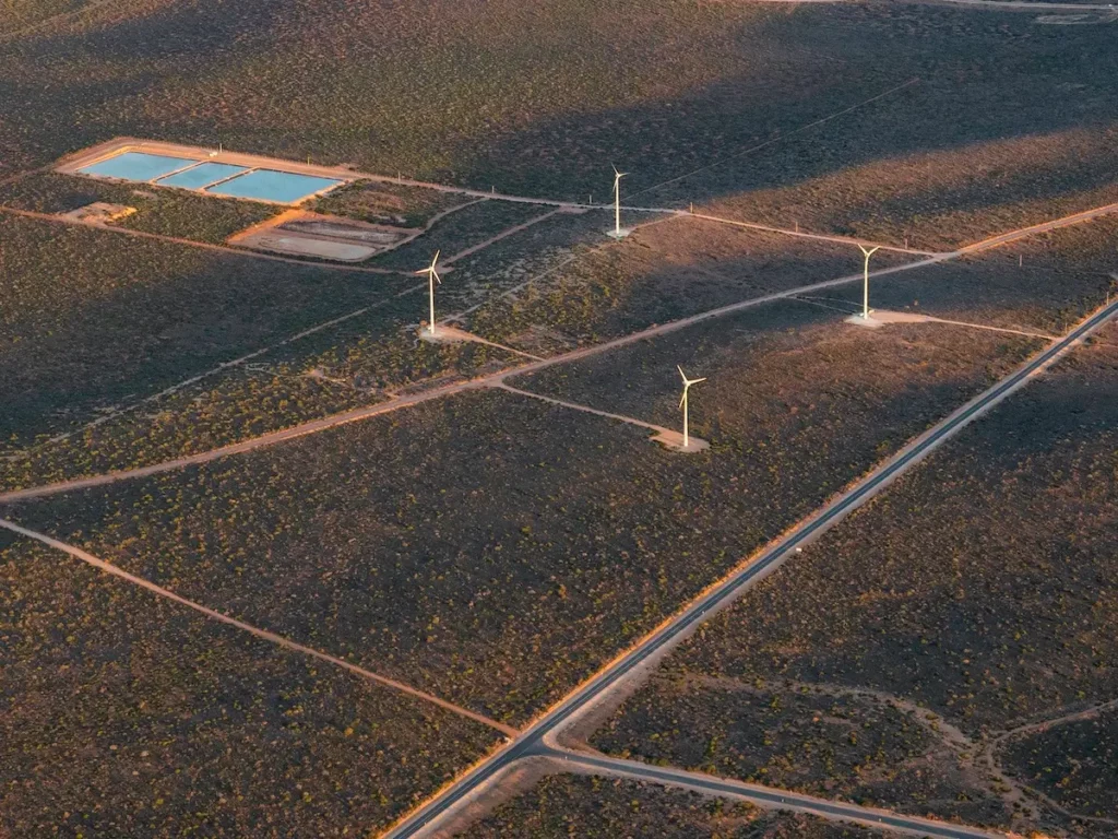 Aerial view of a landscape featuring several wind turbines and two rectangular water reservoirs. The area has sparse vegetation and a grid of dirt roads, creating a pattern across the terrain. Shadows from the turbines are visible on the ground.