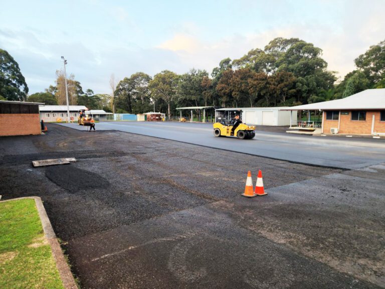 A construction site with workers paving a road. A steamroller is smoothing the asphalt. Traffic cones are placed in the foreground. Buildings and trees are in the background under a partly cloudy sky.