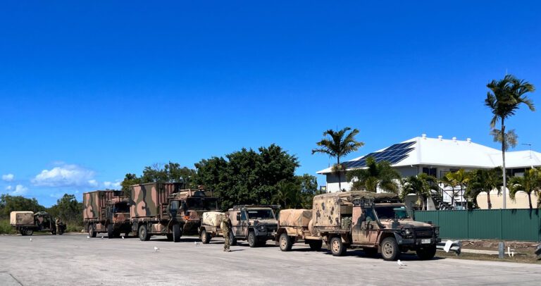 A row of military vehicles parked on a paved area under a clear blue sky. The vehicles are camouflaged, and a couple of palm trees and a white building with solar panels are visible in the background.