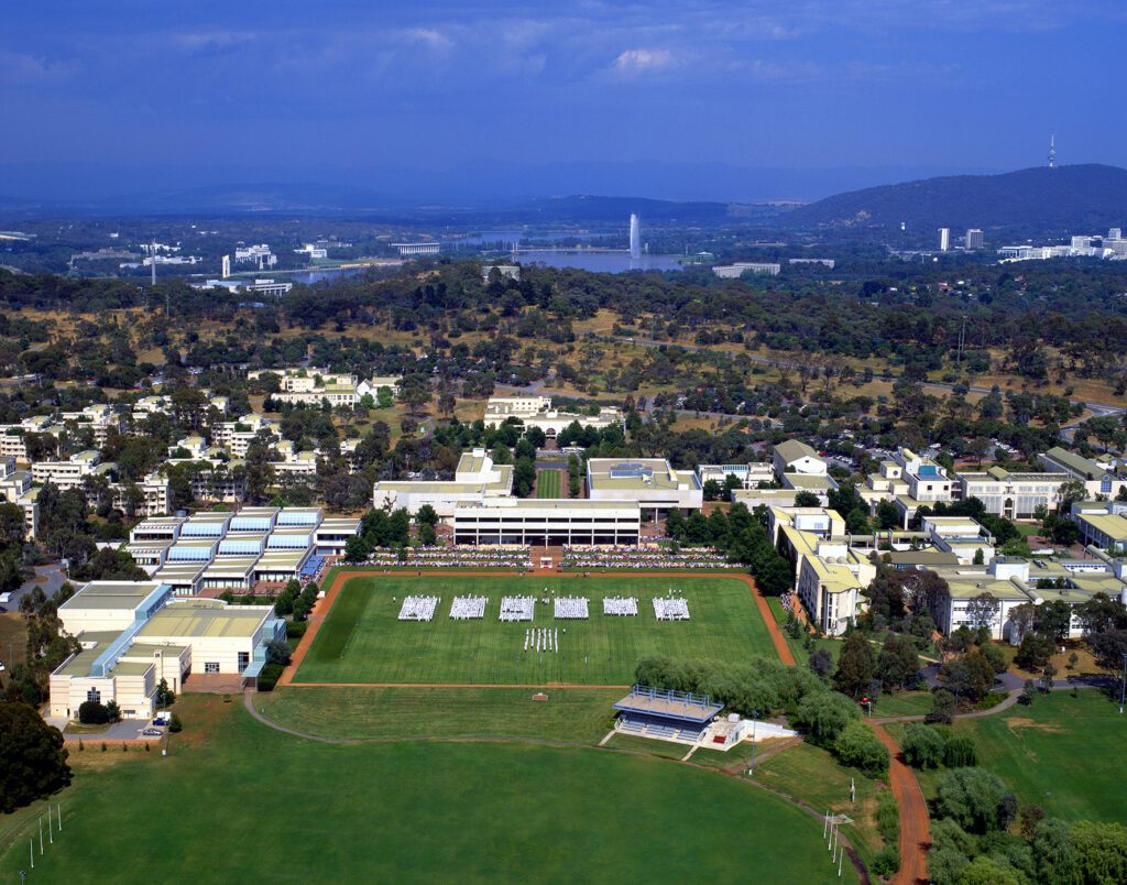 Aerial view of a university campus with multiple buildings surrounded by trees, a large field with white structures, clear blue sky, and distant cityscape visible.