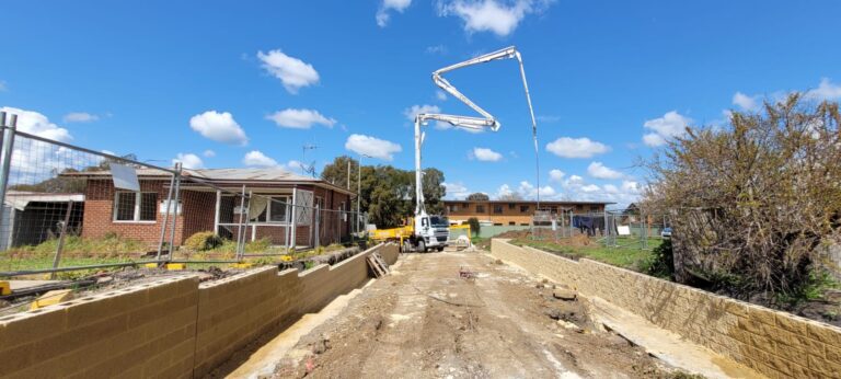 A construction site with a truck and boom crane positioned between two brick walls. A house is on the left, and trees and a building are in the background. The sky is clear with scattered clouds.