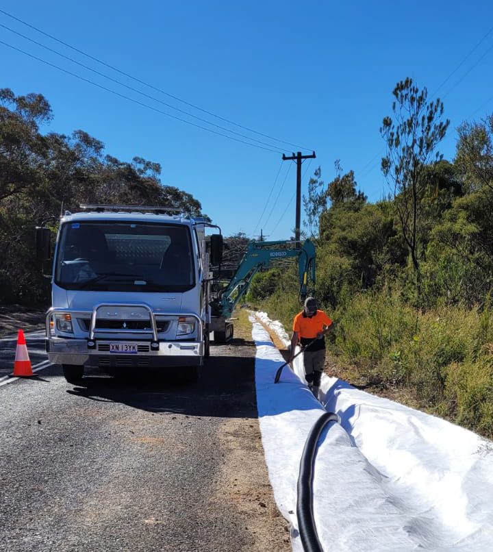 A worker in an orange shirt stands next to a large white sheet on the roadside, guiding a pipe. A small excavator and a truck are parked nearby. Orange cones mark the road under a clear blue sky. Trees and power lines border the scene.