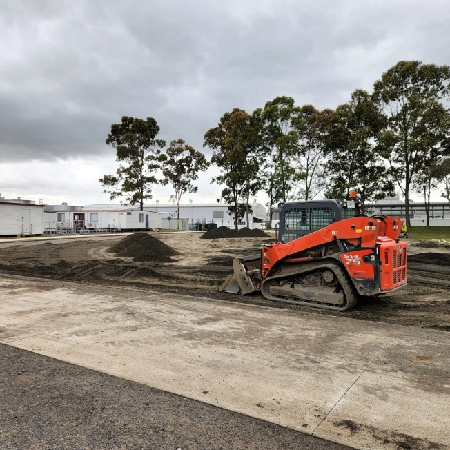 A red skid-steer loader is parked on a construction site with piles of dirt. There are several portable buildings and trees in the background under a cloudy sky.