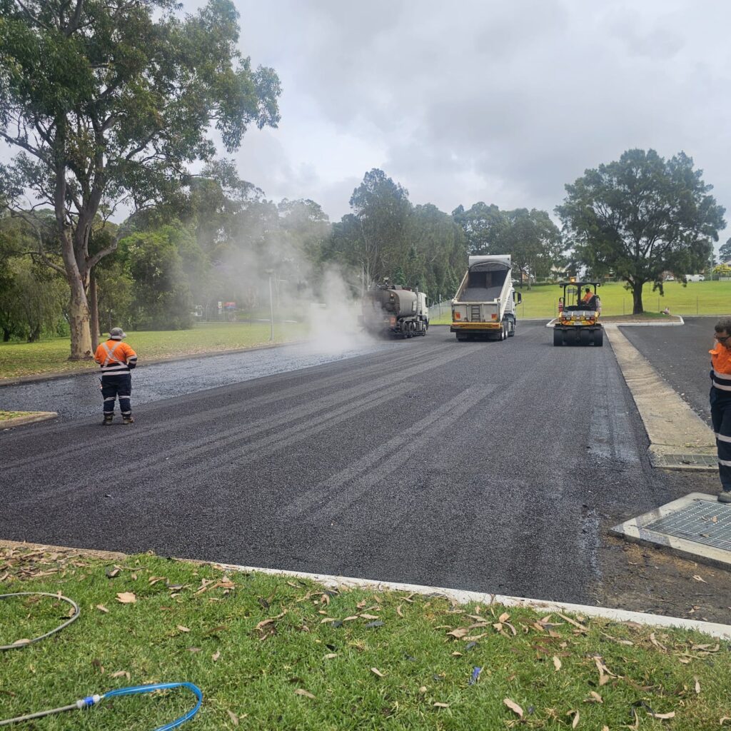 Workers in safety gear are paving a road with asphalt. A truck and a steamroller are on the fresh surface, while steam rises. Trees line the edge of the road, and the sky is overcast.