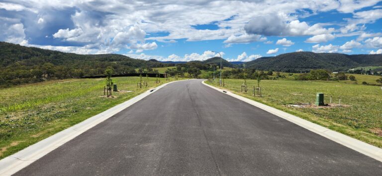 A paved road extends into a lush, green countryside with rolling hills and a partly cloudy sky. Trees and grass line the road, and the horizon features distant mountains under a bright blue sky.