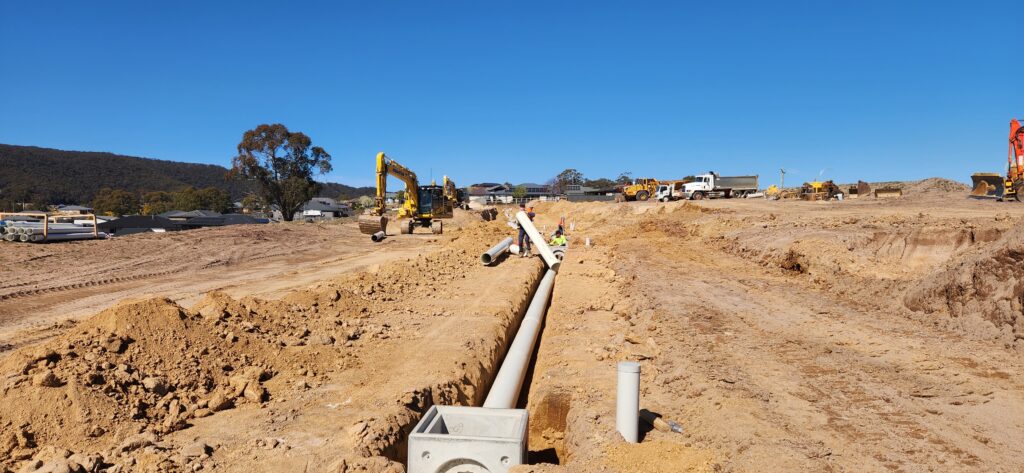 A construction site with workers installing large pipes in a trench. Heavy machinery, including excavators and trucks, are in the background under a clear blue sky. The area is surrounded by dirt piles and sparse vegetation.