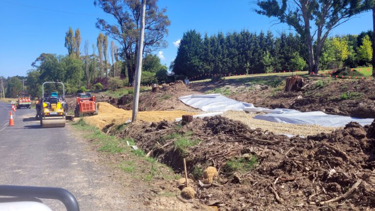 A construction site along a rural road with heavy machinery, including a roller and a digger. The ground is covered with white tarps and piles of gravel. Trees and orange cones line the road, and a clear blue sky is visible overhead.