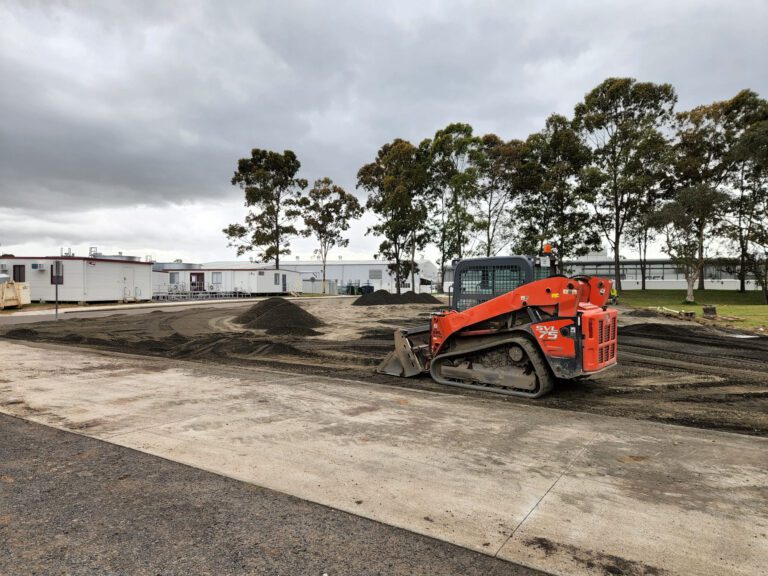 A small orange bulldozer moves soil on a construction site with a cloudy sky above. Trees and several white portable buildings are in the background. The ground is mostly dirt and gravel.