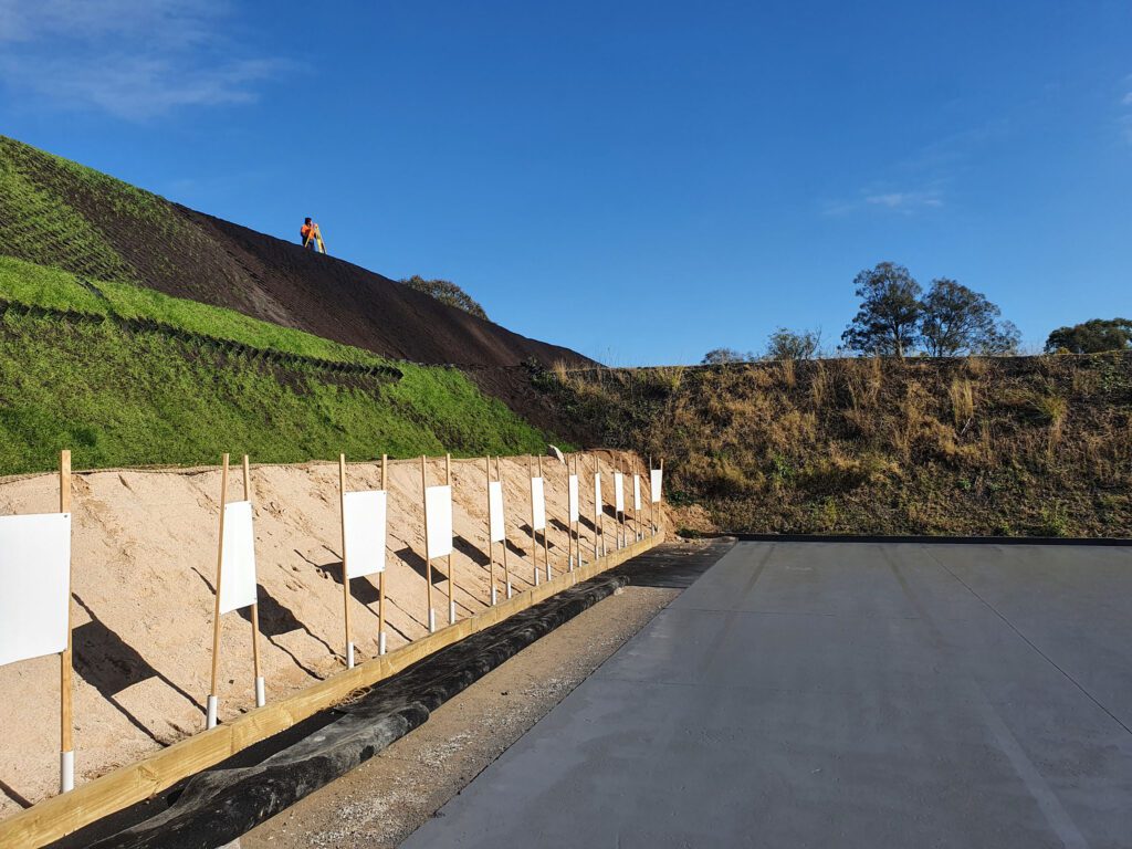 A person in an orange vest works on a grassy slope under a clear blue sky. Below, a series of wooden frames with white panels are lined up on a sandy area next to a paved surface. Sparse trees are visible in the background.