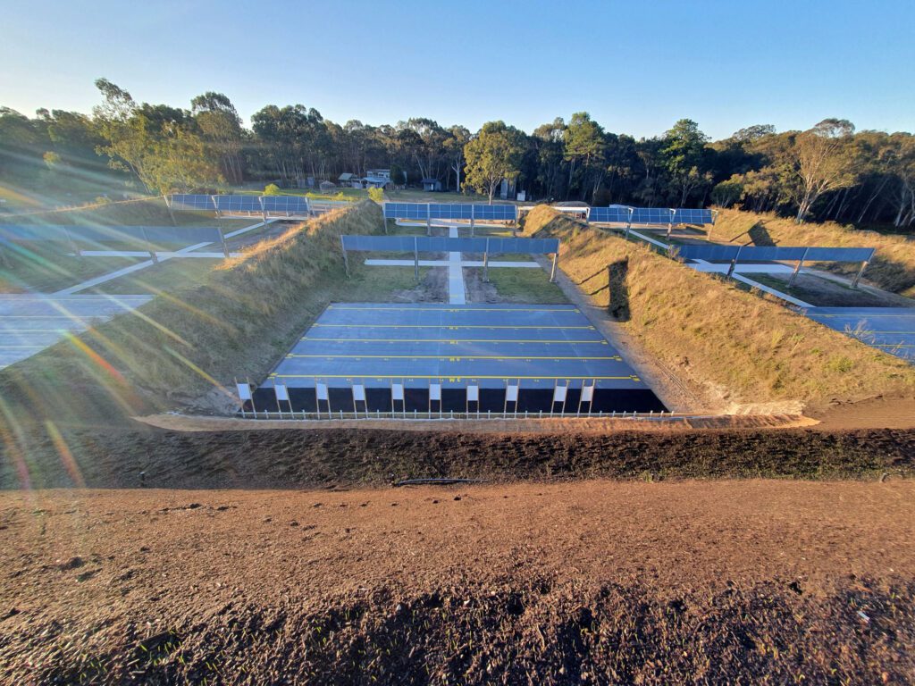 A shooting range with multiple lanes set between grass-covered berms is shown in sunlight. Each lane is separated by earthen walls, and the area is surrounded by tall trees, creating a tranquil outdoor environment.