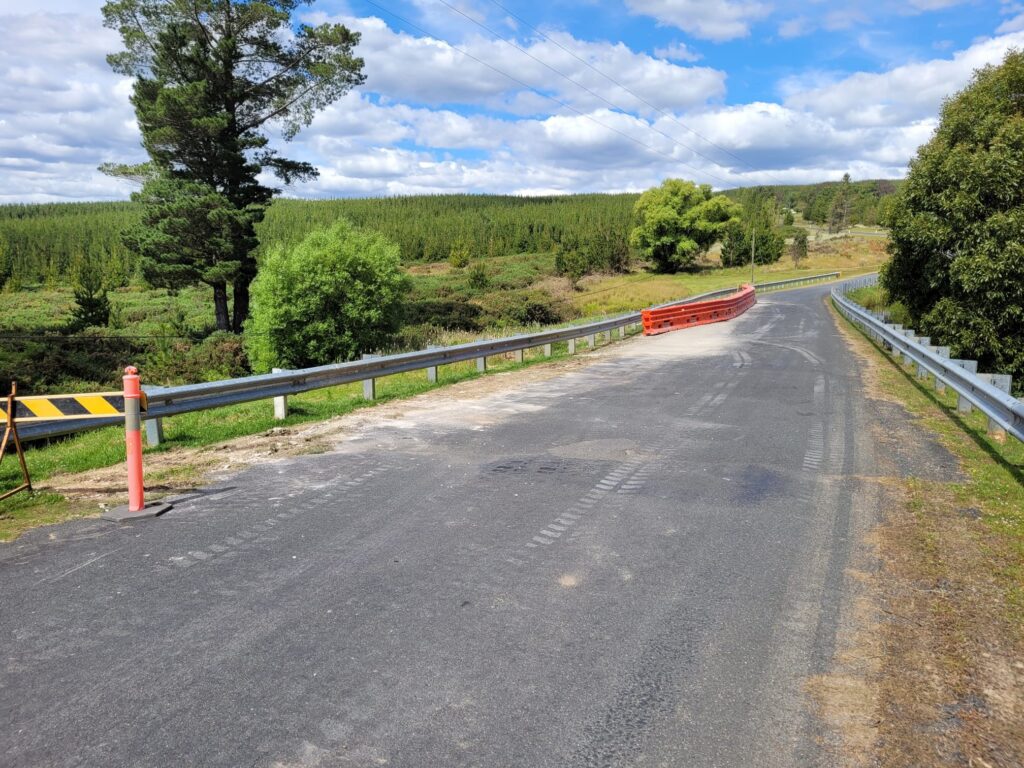 A rural road with guardrails curves through a lush green landscape. A tall tree stands on the left with a forest in the distance under a blue sky with clouds. A small barrier and orange road sign are visible on the roadside.