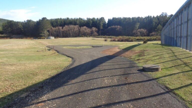 A gravel path curves through a grassy field with scattered trees in the background. The right side shows a metal fence casting shadows on the grass. The sky is clear, suggesting a sunny day in a rural or park setting.