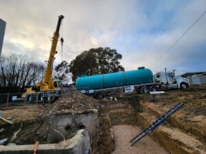 A construction site with a crane lifting a large cylindrical green tank onto a truck. The ground is excavated, revealing soil and debris. Cloudy sky and trees are in the background. A ladder leans against the excavation.