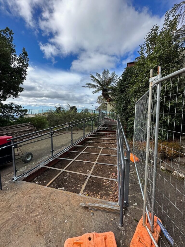 A construction site with metal framework laid on the ground, likely for a pathway or platform. Orange barricades and a temporary fence are on the sides. Trees and a cloudy sky are in the background.