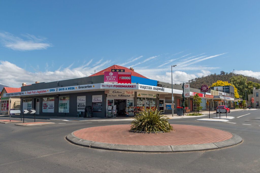 A corner store with signs for a food market and hardware, located in a small shopping area. The building has a red roof and white walls. Trees and a hill are visible in the background under a blue sky with clouds.