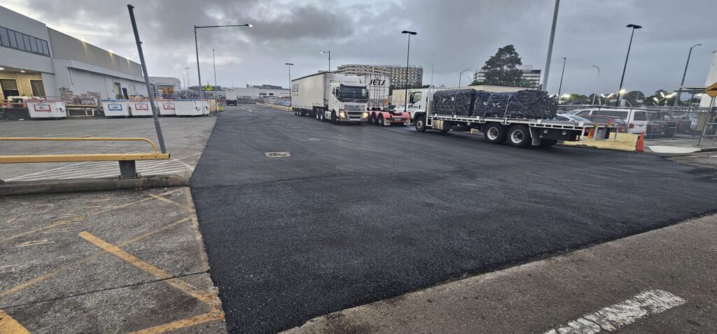 A freshly paved road in an industrial area, with two parked trucks, one with a covered load. The sky is overcast, and there are several streetlights and buildings visible in the background.