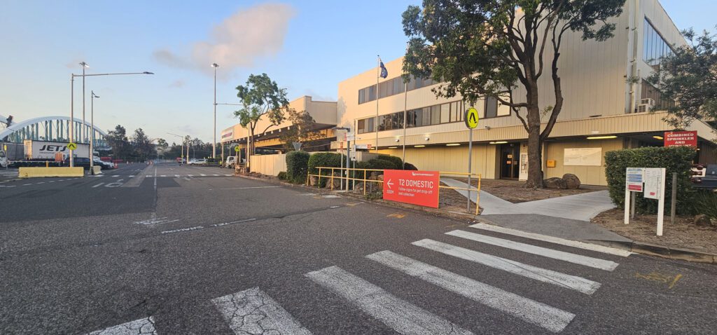 Street view of an airport terminal with a zebra crossing in the foreground. A red sign points towards the terminal entrance. The building is two-story with beige walls and large windows. Trees line the street, and parked cars are visible on the left.