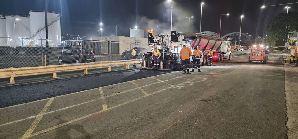 Construction workers in orange safety gear operate heavy machinery at night, resurfacing a road. Bright lights illuminate the site, and industrial tanks are visible in the background.