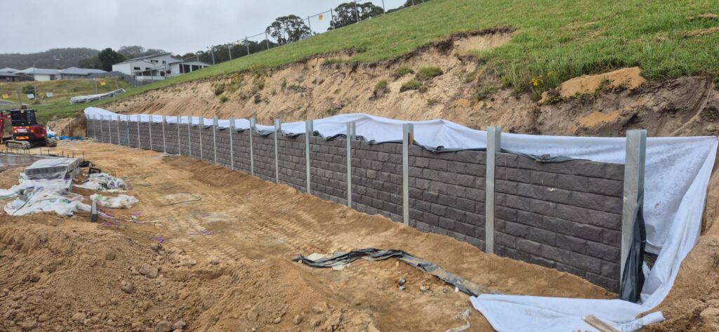 A construction site featuring a newly built retaining wall with gray textured bricks, partially covered with white tarps. The surrounding area shows excavated soil and construction materials. Houses and grassy hills are visible in the background.