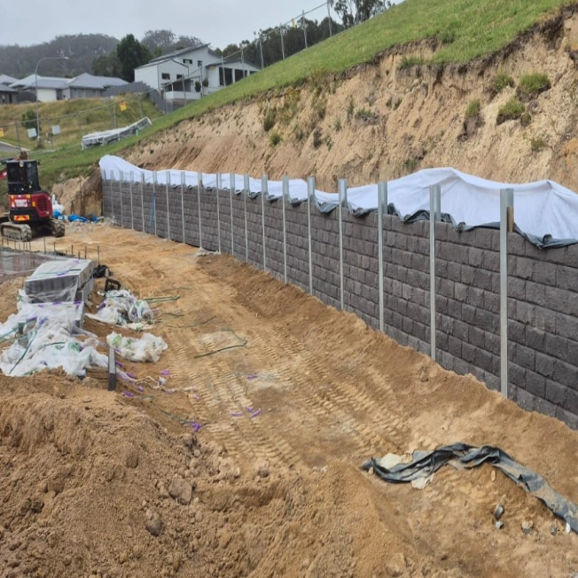 A construction site with a partially completed retaining wall made of concrete blocks. The ground is excavated, and construction materials are scattered nearby. A vehicle is parked in the background, with houses visible on a hill.