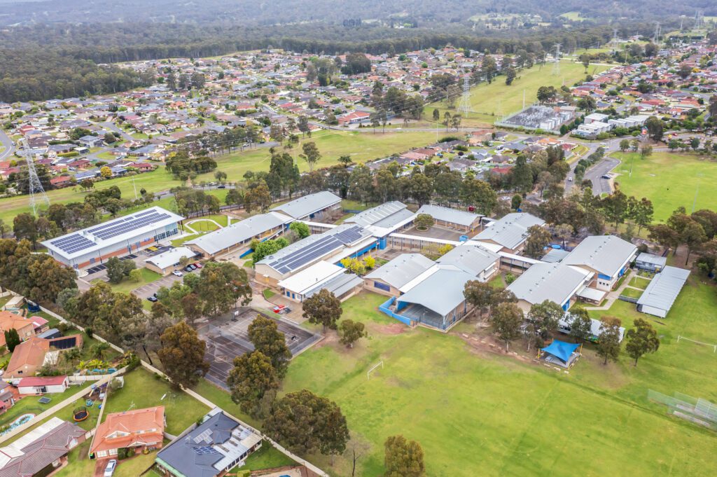 An aerial view of a school complex with several large buildings surrounded by trees and sports fields. Residential houses are visible in the surrounding neighborhood, with a vast expanse of greenery and trees in the distance.