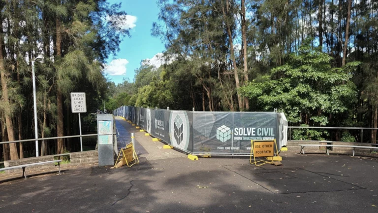 A pedestrian bridge surrounded by trees is closed for construction, with barriers and fencing displaying "Solve Civil" logos. Signs direct people to use an alternate footpath. Bright blue sky with scattered clouds in the background.