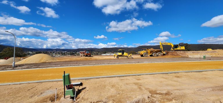 A construction site with heavy machinery, including excavators and bulldozers, working on a dirt landscape. Piles of sand and soil are visible under a bright blue sky with scattered clouds. Mountains are in the background.