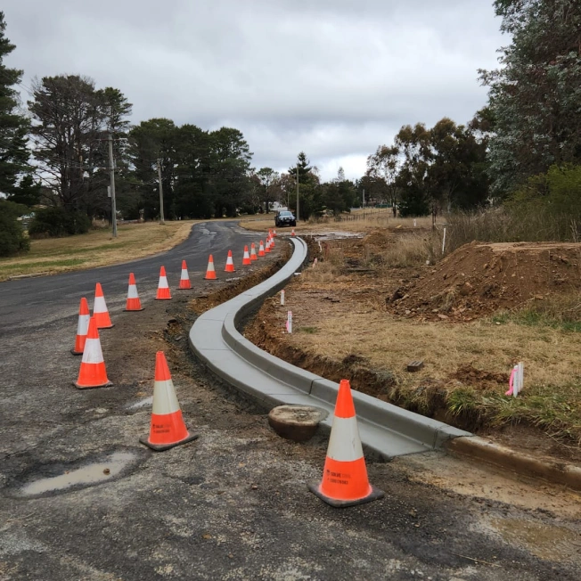 A curved concrete curb under construction along a rural road, lined with orange traffic cones. The surrounding area features grass, dirt, and trees. A vehicle is parked in the distance under a cloudy sky.