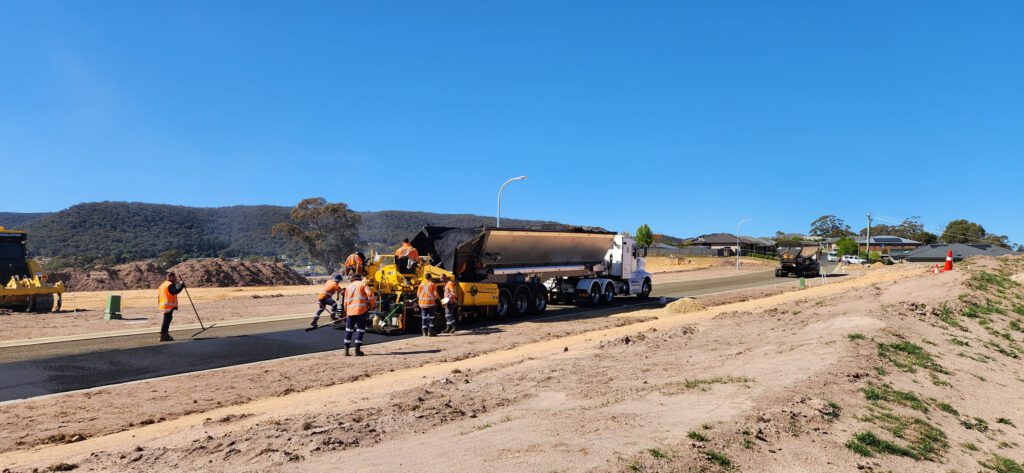 Construction workers are paving a road with heavy machinery under a clear blue sky. A truck carries materials, and trees and hills are visible in the background.