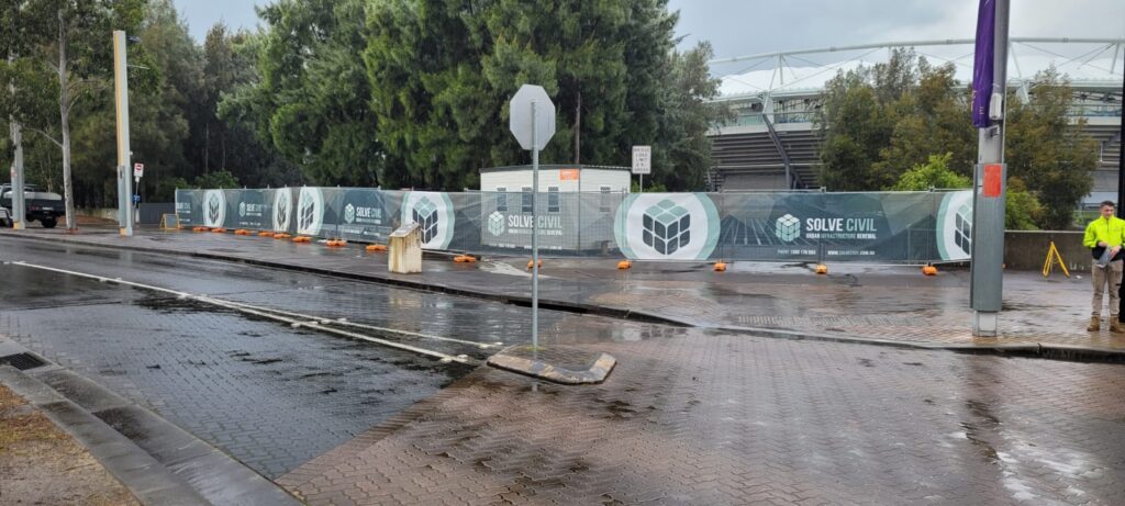 A street view shows a construction site with a fence displaying "SOLVE CIVIL" logos. The wet road and cloudy sky indicate recent rain. Two workers in bright clothing are visible near the fence, along with several parked cars and trees in the background.