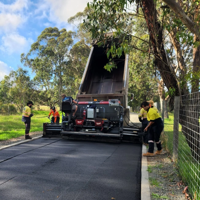 Workers in safety gear operate a machine laying asphalt on a road in a wooded area. A truck with a raised bed unloads material into the paver. Trees and a wire fence line the road under a partly cloudy sky.