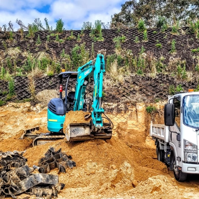 A blue excavator is digging soil on a construction site, with a white dump truck parked nearby. The area has a steep, grass-covered embankment in the background and pieces of black tarp on the ground.