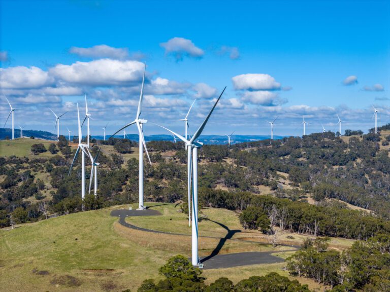 A scenic landscape featuring several wind turbines spread across green rolling hills under a bright blue sky with scattered clouds. The turbines are generating renewable energy, surrounded by lush vegetation and distant hills.